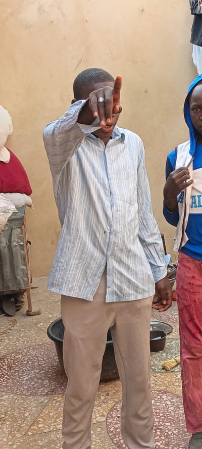 The image features two young individuals in what appears to be an outdoor or semi-outdoor setting in Kaura Goje, Nigeria, under bright daylight.
The central figure is a young person with dark skin and short, dark hair, wearing a light blue and white striped long-sleeved button-up shirt and light brown/khaki trousers. This individual is making a gesture with their right hand, with the index finger pointing upwards, partially obscuring their face. A silver-colored ring with a rectangular setting is visible on their right ring finger. Their mouth is slightly open, possibly indicating speech or a reaction. The light brown trousers have several noticeable damp spots or stains.
To the right, partially visible, is another young person with dark skin, wearing a blue hooded top with white accents and red or pink trousers. Only a portion of their face and upper body is visible. They are looking towards the camera, with their right hand resting near their chest. Their red trousers also display damp spots.
The background consists of a plain, light brown wall. To the far left, a white pillow or plush toy and a red cushion are stacked on a low structure. A corrugated metal object is also partially seen on the left. The ground is covered in a multi-colored mosaic tile pattern, incorporating shades of beige, brown, and white. A black plastic basin sits on the ground between the two people. Closer to the right-hand subject, a small dark pot, a metal tray, and a traditional straw broom are visible on the ground.
The overall scene suggests an informal, possibly domestic or communal environment. No specific activity beyond standing and gesturing is explicitly taking place, though the damp clothing on both individuals might imply recent interaction with water or liquids. There is no clearly legible text visible anywhere in the image.