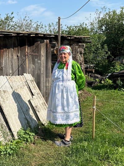 A woman stands in front of a wooden shed in a rural outdoor setting, likely in the village of Sredneye Kechevo, Russia. She is wearing a vibrant green long-sleeved top, a white embroidered apron with intricate lace trim, and a colorful floral headscarf. A black skirt with white polka dots is visible beneath the apron. Her footwear appears to be dark-colored clogs. The shed behind her is constructed of weathered wooden planks and has a simple, aged appearance. To the left of the shed, leaning against it, are several sheets of corrugated material. The ground is covered with green grass, and sunlight casts shadows. In the background, trees and foliage indicate a natural, undeveloped environment. A wooden stake with twine tied around it is visible in the grass to the right of the woman. The overall scene suggests a traditional or rural lifestyle.