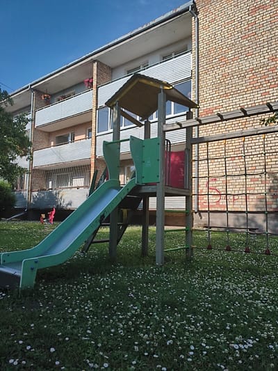 A bright, sunny day is depicted in this outdoor scene set in a residential area of Tukums, Latvia. The foreground features a vibrant green slide and a climbing net that are part of a children's playground structure. The playground is situated on a lush green lawn dotted with white clover flowers. Behind the playground, a multi-story apartment building dominates the background. The building's facade is a mix of light-colored bricks and grey siding, with several balconies visible. Small potted flowers in pink and orange add touches of color to the balconies. A large tree with dense green foliage is visible to the left of the building, providing shade. The sky is a clear, vibrant blue, indicating fair weather. The overall atmosphere is peaceful and suggests a pleasant, warm day, likely during the summer months. No people are visible in the immediate vicinity of the playground, but the presence of the playground equipment suggests it is a place for children to play. Visible text includes "WWW.ANP.LV" printed in white on the side of the slide.