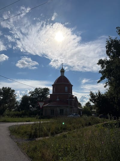 The image shows a brick church with a red dome and a golden cross on top, located in the village of Ryndino, Russia. The sun is shining brightly, partially obscured by scattered clouds. The church is surrounded by trees and vegetation, with a road curving in the foreground. There are also power lines visible in the sky. The scene appears to be peaceful and rural.