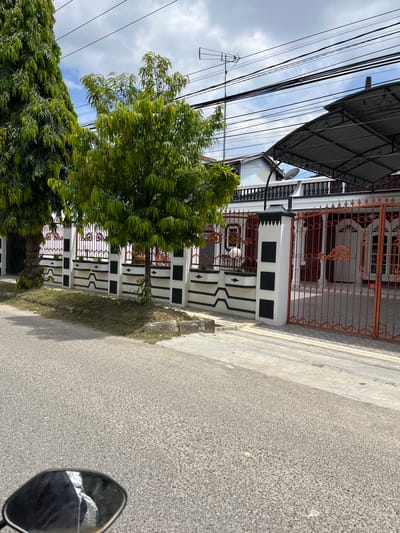 The image captures a street-level view of a property in Langsa, Indonesia, on a partly cloudy day. In the foreground, a textured asphalt road occupies the bottom half of the frame, with a reflective surface, likely a mirror, visible at the very bottom left corner.

The middle ground is dominated by a white and black patterned fence, punctuated by ornate dark spires atop its pillars. Behind the fence, a house with a dark red facade and white window frames is visible. An orange metal gate, featuring intricate scrollwork, stands open. The property is partially shaded by a dark, patterned metal roof structure extending over the gate area. Two trees with dense green foliage are situated to the left of the fence, with one appearing taller and more slender than the other. Electrical wires and a television antenna are prominent in the sky above the house and trees.

The environment suggests a residential area. The overall scene is static, with no visible human activity or interactions. The lighting indicates it is daytime, with shadows cast by the trees and the roof structure.
