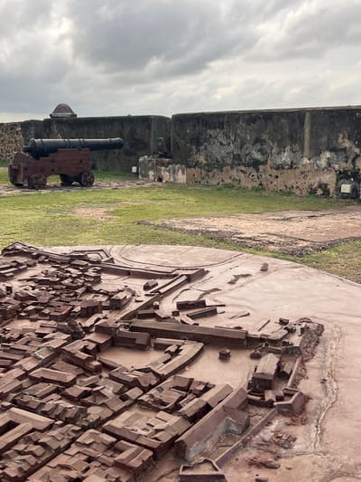 This media file captures an outdoor scene within a historical fort. The primary subjects are a replica of a historical town and a cannon, both placed on grassy terrain. The town replica is a detailed, three-dimensional model made of a reddish-brown material, depicting numerous small buildings arranged in a cityscape. The cannon, made of dark metal and wood, is positioned on a carriage with wheels, resting on stone foundations.
The setting appears to be within the walls of an old fort, indicated by the weathered stone walls visible in the background. The ground is covered with green grass, and parts of the terrain show exposed soil and pathways. The sky is overcast with dramatic, cloudy formations, suggesting either early morning or late afternoon light, and potentially an impending rain shower. The overall atmosphere is one of historical preservation and contemplation.
There is no activity or event taking place; the scene is static, focusing on the display of historical artifacts and the environment. Notable details include the intricate craftsmanship of the town model and the weathered texture of the fort walls, hinting at a rich past. The cloudy sky adds a dramatic element to the scene. No visible text is present in the image. The context provided indicates this location is Galle, Sri Lanka, which aligns with the presence of historical fortifications and the general aesthetic.