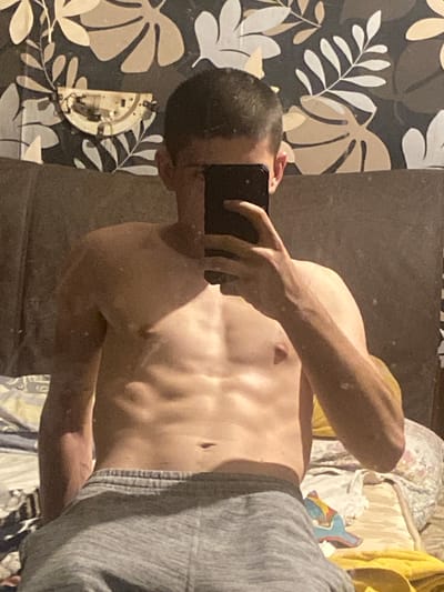 In the city of Gorna Malina, Bulgaria, a young Caucasian male stands shirtless, wearing grey sweatpants. He is positioned in front of a wall adorned with a dark floral wallpaper. The man is holding a smartphone in his right hand, obscuring his face. His physique displays well-defined abdominal muscles. To the left of the man, a circular light fixture is attached to the wall, revealing its internal wiring. The background shows a bed covered with various bedding items, including a patterned quilt and pillows. The lighting in the room is dim, casting shadows and highlighting the contours of the man's body.