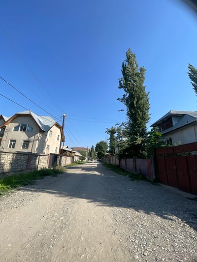 The image depicts a street in the city of Кызыл-Тоо, Kyrgyzstan. The street is unpaved and appears to be made of dirt and gravel, with tire tracks visible. On either side of the street are residential buildings. To the left, a two-story house with a light-colored facade and a metal roof is visible. It has several windows and is bordered by a stone wall. To the right, a similar house with a metal roof and a dark red metal gate is present. Tall, slender trees with dense green foliage are interspersed along the street. Power lines are strung overhead, supported by wooden poles. The sky is clear and blue, suggesting a sunny day. The overall impression is of a quiet, rural or suburban street. There are no people visible in the image, nor are any specific actions taking place.