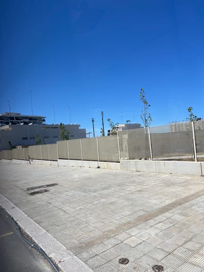 The image, originally oriented vertically and rotated counter-clockwise, depicts a bright, sunlit urban street scene in Bari, Italy. A clear, vibrant blue sky occupies the upper portion of the frame, indicating excellent weather conditions, likely midday or early afternoon given the strong illumination.
Several light-colored, multi-story buildings, possibly residential or office blocks, line the middle ground, stretching horizontally across the view. These structures feature numerous windows and are topped with an array of antennas and satellite dishes. In front of the buildings, a series of white or light-grey, grid-like architectural screens or fences are visible, seemingly separating the buildings from a public walkway. Small trees with green leaves protrude from behind or through these screens, adding touches of nature to the urban environment.
The foreground is dominated by a wide, light-toned paved surface, which appears to be a sidewalk or plaza. The pavement exhibits a varied texture, with some sections composed of smooth, rectangular tiles and others featuring a raised, studded pattern, possibly for tactile guidance. Two round, metallic manhole covers are embedded in the paved area on the right. Further to the right, a grey concrete curb separates the pavement from a darker asphalt road, along which a thick, dark cable runs. No people or vehicles are visible, contributing to a calm, static atmosphere. There is no visible text in the image.