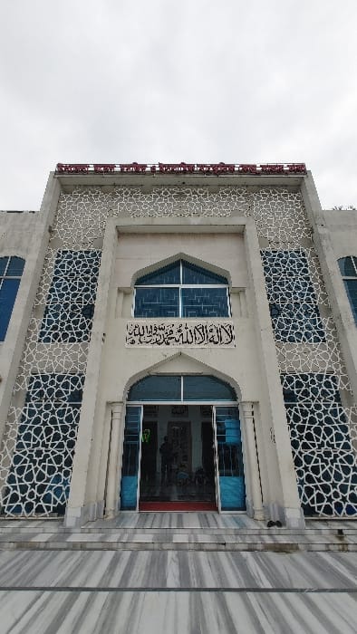 This is a low-angle photograph of the facade of a multi-story building, likely a mosque, associated with Bangladesh Open University, as indicated by a prominent red Bengali inscription reading 'বাংলাদেশ উন্মুক্ত বিশ্ববিদ্যালয়' (Bangladesh Open University) on its upper facade. The structure is situated in the Savar area of Bangladesh.
The building features a light-colored (off-white/light grey) exterior with symmetrical design. Its central section includes an arched main entrance with open, blue-framed glass double-doors. Above the entrance, a rectangular black plaque displays Arabic calligraphy, reading "La ilaha illallah, Muhammadun rasulullah" (There is no god but Allah, Muhammad is His messenger). Directly above this plaque is another arched window with blue-tinted glass.
The facade is extensively decorated with intricate white, abstract geometric patterns that form a screen over blue-tinted windows and wall sections. The building is accessed via wide, horizontally striped grey and white marble steps leading to the entrance. A pair of black shoes are placed on the marble steps to the right of the open doorway. Inside the open entrance, indistinct dark silhouettes of multiple individuals are visible. The sky overhead is uniformly overcast and light grey.