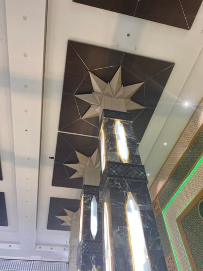 The image depicts the interior of a building, likely a modern architectural space. The perspective is looking upwards towards the ceiling and decorative elements. Dominating the foreground are two large, illuminated columns. These columns have a dark marble-like finish with green and black checkered patterns at their bases and mid-sections. Each column features a tall, narrow, arch-shaped opening illuminated from within, casting a bright white light.

Above the columns, the ceiling features dark brown rectangular panels with geometric patterns created by thin metallic lines. Emerging from these panels are three-dimensional starburst or faceted geometric sculptures, rendered in a light wood or stone material.

To the right of the columns, a wall is decorated with intricate geometric tiling. This tiling features a repeating pattern of small squares and octagonal shapes in shades of beige, green, and red. A bright green neon strip light runs diagonally across this patterned wall.

The ceiling itself is white and appears to be a suspended ceiling with recessed circular lights visible in some areas. The overall impression is one of a contemporary and possibly luxurious interior design. There are no people visible in the image. The scene does not provide any direct visual cues to pinpoint its location to Aluekambuk, Indonesia, or any other specific city.