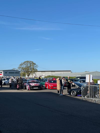 This image depicts an outdoor scene in what appears to be a parking area adjacent to buildings and a street. The location is Silverstone, United Kingdom, based on a visible vehicle registration plate.
The environment features a clear blue sky, a tree with sparse foliage, and buildings of light color. A row of parked cars lines the area, including a white van, cars of various colors (black, red, green), and a black car with anime-style decorative elements.
People are present, some standing next to or near the cars. Interactions are not clearly discernible, but some may be socializing or observing the vehicles.
The scene appears to be a car meet or a gathering of automotive enthusiasts, possibly related to an event at the Silverstone circuit.