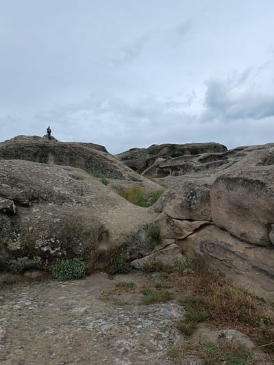 The image depicts the ancient cave city of Uplistsikhe, Georgia, characterized by its unique rock-hewn architecture and natural landscape. In the foreground, the terrain is dominated by large, weathered rock formations with varied textures and colors, ranging from light gray to tan. Patches of dry grass and sparse green vegetation grow in the crevices and on the surfaces of the rocks.
In the middle ground, the scene opens up to reveal more complex rock structures, including carved niches and hollows, indicative of human habitation. The sky above is overcast with a layer of gray clouds, suggesting a diffused, natural light.
In the background, a solitary figure, appearing to be a person, stands on a prominent rock outcrop on the left side of the image. The figure is small in scale relative to the surrounding landscape, emphasizing the vastness of the ancient site. The overall impression is one of a serene, historical location with a rugged natural beauty.