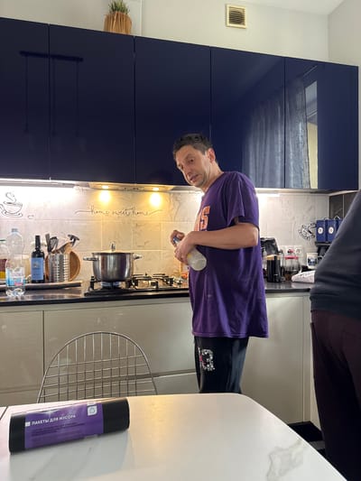 In the city of Ptichnoye, Russia, a man is standing in a kitchen, facing left with his head turned towards the camera. He is wearing a purple t-shirt with orange lettering and black pants with a white floral design. He is holding a bottle in his right hand and appears to be pouring its contents into a pot on a stovetop. A metal pot with a lid is on a gas burner, which is lit. To the left of the pot are a clear plastic bottle of water and a dark glass bottle. Behind the stovetop is a tiled backsplash with the words "home sweet home" scripted in black. The kitchen features dark blue cabinets above and white cabinets below the countertop. A metal chair is partially visible in the foreground. A roll of black trash bags with purple labeling is on the white table in front of the chair. The overall lighting suggests it is daytime.