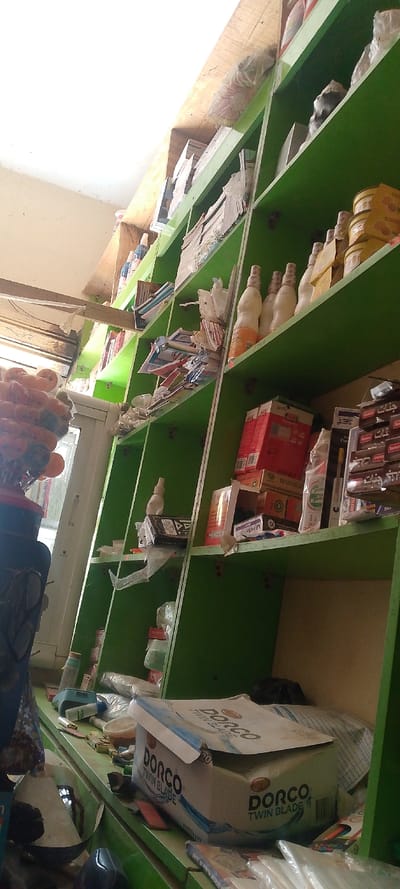 The image depicts the interior of a small retail establishment or storage room in Danbare, Nigeria, characterized by a dominant green multi-tiered shelving unit extending from the foreground to the background. No individuals are present.

The shelving unit is filled with various merchandise. On the upper shelves, multiple white plastic bottles with orange-colored caps, stacks of paper or notebooks, and small yellow-lidded cylindrical containers are visible. Clear plastic bags containing unidentified items are also stacked. Mid-level shelves display more stacks of paper, small white spray bottles, red and brown cardboard boxes, and other packaged goods, some partially obscured by plastic sheeting.

The lower shelf or countertop area features a prominent white cardboard box labeled "DORCO TWIN BLADE" multiple times, suggesting shaving razors. Adjacent to this box are a small light-blue digital scale, several additional plastic bottles, and various other packaged items. To the far left, a dark blue/black container with a handle is partially visible.

In the upper-left quadrant of the image, a white-framed doorway is present, partially open. A transparent plastic bag containing numerous spherical, orange-colored objects hangs near the doorway. Above the doorway, a rough wooden beam spans horizontally. The ceiling is white, and portions of the wall visible on the left are an off-white or cream color. The scene is static; no actions or interactions are depicted.