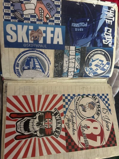 The image shows an open notebook filled with stickers. The notebook is aged and appears to be a scrapbook or journal.
In the top half of the image, there are several stickers. One prominent sticker features the text "SKIFFAPAUL" in large blue letters, with a small octopus placed over the letter "I". Below this, the text "@SKIFFAPAUL" is visible. To the right of this, a sticker depicts a police helmet with the German word "POLIZEI" (police) reversed on the visor, and text "1312" and "HATE COPS" are also present. Other stickers in this section include circular emblems with German text and sports-related imagery.
The bottom half of the image features more stickers. One large sticker on the left shows a skull with fangs and hands making a rock-and-roll gesture, with a red and white striped background. The skull has a flag wrapped around its head with a cross symbol on it. To the right, a collection of circular stickers and other designs are visible. One circular sticker has text that includes "GELSENKIRCHEN ULTRAS". Another circular emblem prominently displays the number "87" in a bold red font, with German text above and below it. The phrase "15 Years" is also visible in stylized lettering. The city of Gelsenkirchen, Germany, is mentioned in text on one of the stickers, likely indicating a connection to a football club or fan group from that location. The environment appears to be an indoor setting, likely a room or desk where the notebook is placed.