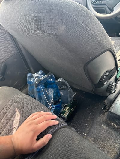 This is a medium shot, eye-level media file taken outdoors in Opatów, Poland. The scene shows the interior of a vehicle, likely a car, with a focus on the passenger seat. A person's hand is resting on the seat, indicating a human presence.
On the floor of the vehicle, there is a plastic-wrapped package containing several blue bottles of water. Next to the water bottles, there is a green and silver can, possibly a beverage can, lying on its side. The floor of the car is covered in a dark carpet which appears to have some debris on it.
The seats are upholstered in a dark gray fabric with a textured pattern. Part of the steering wheel and dashboard are visible in the upper left corner of the image. The overall impression is one of a casual setting, perhaps during a road trip or a stop. There are no strong indicators of the time of day or weather conditions, but the lighting suggests it is daytime. The visible text on the can is partially obscured, but some lettering is discernible.