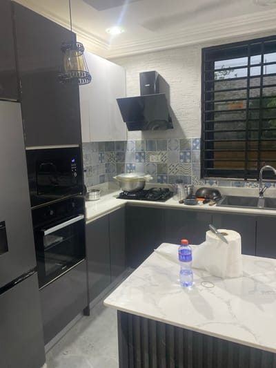 The image displays a modern kitchen, presented sideways as if rotated 90 degrees counter-clockwise. The space features sleek, dark grey lower cabinets and built-in appliances, paired with white upper cabinets. The countertops are a light color, with a prominent kitchen island or peninsula featuring a white marble-effect surface.
A decorative blue and white patterned tile backsplash extends above the gas stovetop, over which a black range hood is mounted. A pot rests on the stovetop burners. To the left, integrated black appliances, including an oven and another unit (possibly a microwave), are built into the cabinetry. A window with black security bars is visible, providing natural light, and a stainless steel sink with a faucet is positioned beneath it.
On the kitchen island/peninsula, a roll of paper towels with a knife stuck in it, a plastic water bottle, and a rubber band are present. The kitchen is illuminated by both recessed ceiling lights and a decorative pendant light with a dark metal cage and an amber-toned inner glass, hanging over a section of the countertop. The walls have a light, textured finish, and crown molding is visible along the ceiling line. Electrical outlets are present on the backsplash. No individuals are visible, and no discernible text is legible other than general appliance branding.