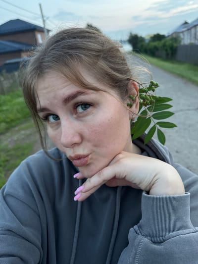 A young woman with light brown hair, casually dressed in a grey hooded sweatshirt, is captured in a close-up, outdoor selfie. She has her hand resting on her chin, and a branch with green berries is tucked behind her right ear. Her gaze is directed towards the camera, and her lips are pursed in a kissing gesture. The background suggests a semi-rural environment, with a paved path, greenery, and hints of residential buildings and a body of water, consistent with a location in the city of Gorniy, Russia. The lighting indicates it is either early morning or late evening, with a cloudy sky.