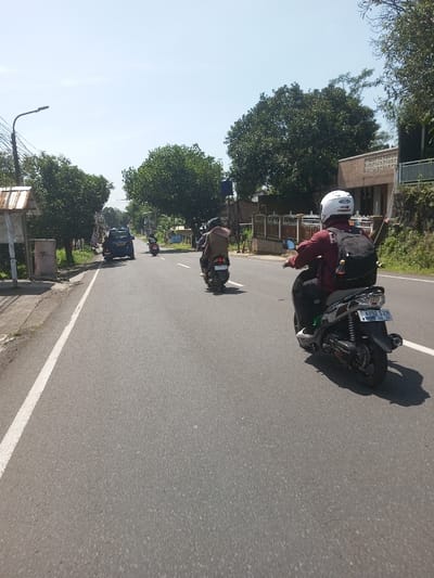 The image displays a daytime street scene on an asphalt road in Beteng_Pangkhar_Somthrang, Bhutan, under a clear, bright blue sky. In the immediate foreground, a person is visible from behind, operating a dark gray/black scooter. This individual wears a white full-face helmet, a maroon long-sleeved shirt, dark pants, and white athletic shoes, carrying a black backpack from which a brown bottle hangs. The scooter's license plate, partially visible, reads 'B 6492 SXN' with '08-28' below it. Ahead and slightly to the left, a second person, also viewed from behind, rides a dark scooter, wearing a light brown jacket and a dark helmet. Further down the road, a blue SUV or compact car is visible on the right side.
The left side of the road features a utility pole supporting a street light, a small open-sided structure with a corrugated metal roof, and an abundance of dense green foliage and trees. The right side of the road is bordered by a concrete fence with dark brown vertical slats, behind which stands a light brown multi-story building with a distinctive patterned concrete railing on an upper level. Beyond the building, a vegetated slope rises, covered in more trees. Shadows cast by the vehicles and roadside objects indicate direct sunlight. The scene depicts typical road traffic, with multiple vehicles traveling forward on the paved surface.