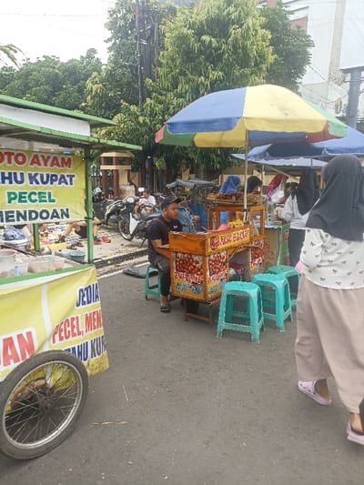 The image shows a street food vendor in Purwokerto, Indonesia, located at latitude: -7.417, longitude: 109.249. The vendor is selling a variety of street food, including what appears to be fried snacks. The vendor is a man in a black shirt and black pants who is sitting on a stool and facing the camera. He is attending to his food stand. There is a green cart with a yellow sign displaying the vendor's food options and a blue umbrella providing shade for the customer. The customer is a woman in a cream-colored dress, white and black patterned top, and hijab. She is walking away from the food stand, carrying a bag. There are two other people on the sidewalk. One is a woman in a blue shirt and black pants standing behind the food stand. There is also a man sitting on a motorcycle nearby. The street is paved with grey asphalt and lined with trees on either side. A brick wall is visible in the background, as well as a building with a white facade. The environment is bustling and characterized by street food activity.