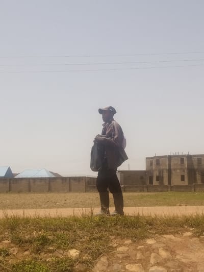 A lone individual, a man wearing a baseball cap and a jacket over a bag, walks along a dirt path in Reifild-Ist, Nigeria. The man is dressed in dark clothing and appears to be carrying a dark bag over his shoulder. Behind him, a fence separates a dusty, open area from structures that appear to be under construction. The sky is overcast and a pale blue, with visible power lines stretching across the frame. The foreground consists of dry, patchy grass and dirt. The scene suggests a rural or semi-urban environment within the specified location.