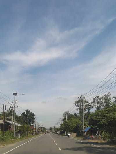 The image depicts a rural road in Jurong Pandee, Indonesia, on a bright, partly cloudy day. The road, paved with asphalt, is marked with white dashed lines and a solid white line on the left shoulder. Several utility poles with numerous power lines stretch along both sides of the road. On the left side, houses with tin roofs and vegetation are visible. On the right side, there is a small blue-roofed structure constructed from wood and bamboo, with some items stored inside. The road is lined with trees and lush green foliage. Two motorcycles are visible on the road, one moving away from the camera on the right and another approaching on the left. The sky is a light blue with scattered white clouds.