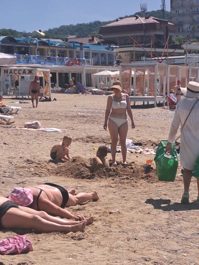 On a sunny day at the Dzhubga beach in Russia, several individuals are engaged in various beach activities. In the foreground, two individuals are lying prone on the sand. One is wearing a black swimsuit and a pink checkered sun hat, while the other is wearing a black swimsuit. Further back, a child is sitting in the sand, seemingly playing. Another child is partially buried in the sand behind him. A woman in a white bikini and a sun hat stands nearby, looking towards the right. To her right, a person wearing a white cover-up and a straw hat walks away from the camera, carrying a large green bag. In the background, a strip of buildings line the coast, with tents and beach chairs set up on the sand. The overall scene depicts a typical summer day at a coastal resort.