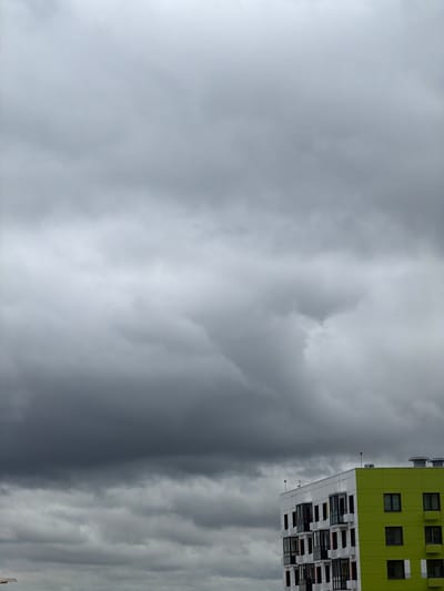 This media file depicts a building against a cloudy sky. The building appears to be modern, with a distinctive architectural style. It has a bright green section on the upper floors and a white section below, featuring numerous windows and balconies arranged in a geometric pattern. The sky is filled with dramatic, gray clouds, suggesting overcast or stormy weather. The lighting is diffused, characteristic of a cloudy day. The orientation of the image is such that the building is shown vertically, with the sky occupying the majority of the frame. There are no people visible in the image. The image captures a moment of natural atmospheric conditions juxtaposed with man-made structure. The overall mood conveyed is somewhat somber due to the heavy cloud cover. There is no visible text in the image.