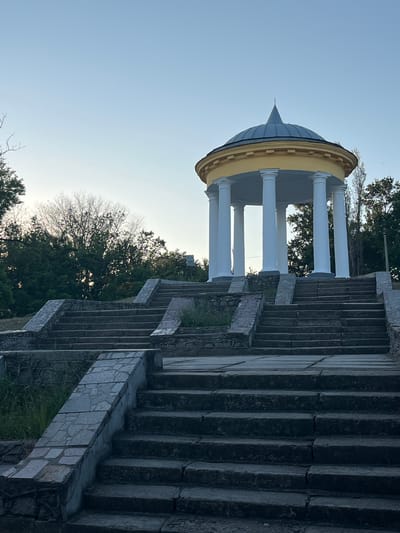The image depicts a grand, open-air pavilion with a domed roof and white columns, set against a clear, pale blue sky. The pavilion is situated at the top of a wide, stone staircase. The staircase is constructed from large, dark grey stone blocks and features decorative stonework along its edges. Trees with dark green foliage and some bare branches surround the pavilion and staircase, suggesting a temperate climate and possibly the transition between seasons. The lighting indicates it is daytime, likely morning or late afternoon, due to the soft, natural light. There is no visible text in the image. The overall impression is one of architectural grandeur and serene natural beauty, suggesting a public park or historical site in Voznesensk, Ukraine.