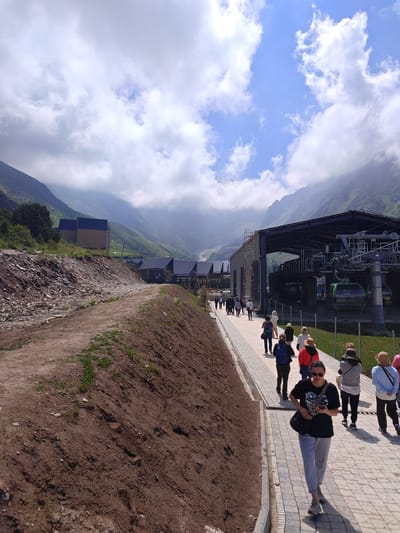 A group of people are walking along a paved pathway in a mountainous area. The pathway is lined with a raised dirt embankment on one side and a fence on the other. In the background, there is a large building with a metal roof, likely a cable car station, with several green cable cars visible. Beyond the building, more structures, possibly houses or cabins, are nestled into the green, rolling hills. The mountains in the distance are shrouded in clouds, giving the scene a dramatic atmosphere. The sky is a mix of blue with patches of white clouds, suggesting it is daytime, likely late morning or early afternoon. The lighting indicates sunny weather with some cloud cover. The people are dressed casually, some wearing hats, and appear to be tourists exploring the area. The overall mood is relaxed and observational. No visible text is present in the image.