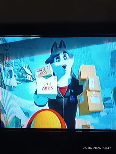 The media file shows an animated husky dog, likely a character from a children's show, holding several cardboard boxes. The husky is wearing a dark jacket over a red shirt. The background depicts a snowy, mountainous scene with houses resembling a village, suggesting a winter setting. The overall atmosphere appears cheerful, with the husky's wide eyes and open mouth conveying a sense of surprise or excitement. The boxes the husky holds have text that appears to be "ABDS" on them, possibly indicating a delivery service or product. The lower right corner of the image displays a timestamp: "25.06.2026 23:47". The image resolution is low, with visible scan lines and pixelation, suggesting it was captured from a screen. The location context is Bolhrad, Ukraine.