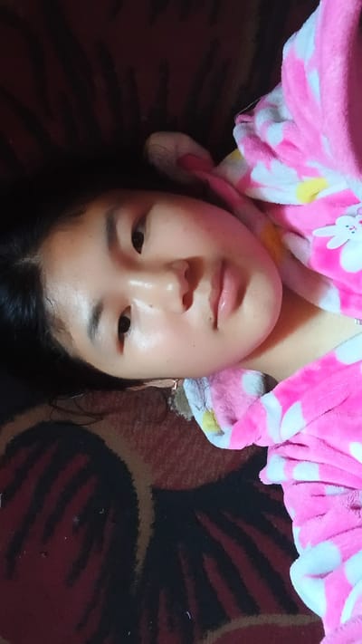 A young East Asian female lies on her back, looking upwards at the camera. She has dark hair and is wearing a pink and white polka-dot robe with a hood. The background appears to be a carpet with a dark red and black patterned design. The overall lighting suggests an indoor setting, possibly daytime due to the natural light illuminating her face. The image is taken from a slightly elevated angle, focusing on her face. The location is identified as Uchkun, Kyrgyzstan.