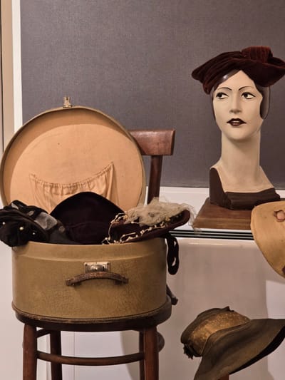 The image depicts a collection of vintage hats and a mannequin head, displayed indoors. The primary focus is on a large, round, light brown hatbox, open to reveal several hats inside. One hat appears dark velvet with pearl embellishments. Another is a dark velvet cloche hat. A third hat is partially visible with delicate lace trim and pearls. The hatbox rests on a wooden stool with a curved backrest, positioned against a textured grey wall.
To the right of the hatbox, a mannequin head wearing a dark brown velvet turban is visible. The mannequin head is light-skinned with painted features, including dark eyeliner and lipstick. It stands on a wooden base, which is placed on a white shelf. Next to the mannequin head, a portion of another hat, possibly straw, is visible on the far right. In the foreground and to the right of the stool, another dark-brimmed hat with a textured band is displayed. The lighting suggests an indoor setting, likely a museum, antique shop, or personal collection. There is no visible indication of the location being in the city of Lom, Bulgaria.