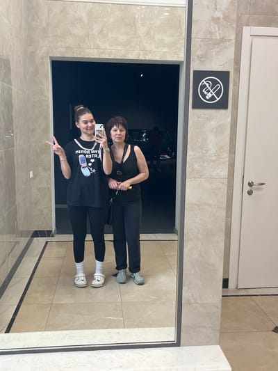 In Verkhnyaya Pyshma, Russia, two individuals, an adult female and a young female, are captured in a mirror selfie. The young female, on the left, is wearing a black t-shirt with a graphic of a flip phone and hearts, black leggings, and white Crocs. She is making a peace sign with her right hand. The adult female, on the right, is wearing a black tank top and black pants, holding a yellow object and a black purse. Both are standing in what appears to be a hallway with marble-tiled floors and walls. A "no smoking" sign is visible on the wall to the right of the mirror. The reflection in the mirror also shows a dark opening, possibly an entrance to another area, with a car and people visible within.