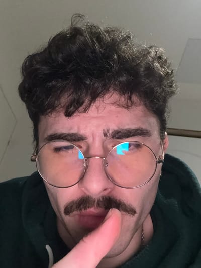 A man with dark, curly hair wears round glasses. He appears to have drawn thick eyebrows that nearly connect across his forehead. A similar marking is drawn above his lip, resembling a mustache. His skin tone is fair, and he wears a dark green hoodie. A finger touches his lip, partially obscuring the fake mustache. The background is a plain white or light-colored wall.
