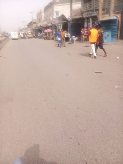 The image captures a daytime street scene, likely in a market or commercial district, characterized by a hazy atmosphere. The primary focus is a wide asphalt road, which occupies the majority of the foreground and midground, stretching into the distance.
In the immediate foreground, the road surface is visible, appearing to be paved with asphalt or similar material, showing some wear and scattered small debris. A shadow, likely cast by the photographer or a nearby object, is partially visible in the bottom left corner. Towards the right side of the road, there's a dark, flat object that resembles a flip-flop or sandal lying on the ground, and a small, light-colored object further to its right, possibly a piece of fruit or trash.
In the midground, on the right side of the road, several people are visible, walking or standing near the roadside. Three men are walking towards the left of the frame. One man on the far right wears dark shorts and a patterned short-sleeved top, with pink flip-flops on his feet. Another man, walking beside him, wears an bright orange or yellow vest over a white long-sleeved top and light-colored trousers, with dark sandals. Further to their left, another man in a blue and white striped shirt and light-colored trousers is pushing what appears to be a small, wheeled cart or trolley. To his right, another man stands, dressed in a light brown or orange long-sleeved shirt and trousers.
Along the right side of the road, there's a row of shops or market stalls. These structures vary in appearance; some seem to be simple wooden or metal stands with tarpaulin or fabric coverings, while others are more permanent buildings. Merchandise, including clothing items hanging outside some shops, is discernible. Notably, several large solar panels are displayed prominently outside some of these establishments, suggesting a business dealing in solar power equipment. One stall has a reddish-brown umbrella providing shade.
In the background, the road continues, with several vehicles, including cars, vans, and possibly auto-rickshaws, driving or parked. On the far left, a white vehicle, possibly a truck or large van, with stacked items covered in white sacks, is moving away from the camera. The buildings lining the street in the background appear to be multi-story and are somewhat indistinct due to the haze and distance. Power lines and utility poles are visible above the buildings. The overall impression is that of a bustling street in a densely populated area. The hazy conditions suggest either dust in the air or perhaps early morning/late afternoon light.