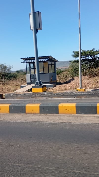 A gray and glass toll booth sits on a concrete platform with a ramp. It has a slanted roof with a blue tint. The base of the supports is painted yellow. A utility box is attached to a gray pole on the left. Another gray pole stands to the right of the booth. The concrete curb fronting the booth has alternating yellow and gray stripes. A gray paved road fills the foreground. Sparse vegetation and distant hills are visible. The sky is clear blue.