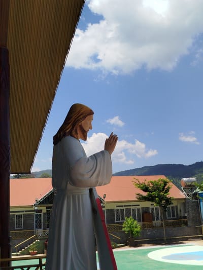 A statue of Jesus is positioned in the foreground, facing right and slightly away from the viewer. The statue is white, with Jesus depicted with long brown hair and a beard, wearing a flowing white robe. His right hand is raised, with the palm facing outward and a stigmata visible on the palm. In the background, a building with an orange roof is visible, along with green trees and rolling hills. The sky is blue with scattered white clouds. The scene appears to be outdoors, possibly at a religious site or community center. The image quality is moderate, with some graininess and minor artifacts. The lighting suggests it is daytime, with the sun casting shadows. The statue is life-sized or larger. The building in the background has a modern architectural style with a tiled roof and windows. There is a green court or pavement in the foreground. The overall atmosphere is peaceful and serene.