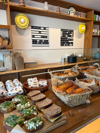 This image depicts the interior of a cafe in Santa Eulària des Riu, Spain. The scene is viewed from a slightly elevated angle, looking down onto a wooden counter displaying various food items and cafe signage.
On the counter, in the foreground, are several open-faced sandwiches on dark bread, topped with avocado, cheese, and greens. Behind these, a terracotta dish holds individually wrapped pastries or sandwiches. Further back, a large wicker basket overflows with golden-brown croissants. Another basket next to it contains more pastries. To the left, a stack of disposable brown paper containers is visible.
On the wall behind the counter, two black letter boards are mounted. The left board displays "LUNCHBOX 16 OR BY WEIGHT LETS EAT REAL LETS EAT JUNTOS." The right board lists menu items and prices: "SHROOMIES COFFEE 4 FOCUS LATTE 4 CHAGACCINO 4 GLOW LATTE 4 HOMEMADE PISTACHIO LATTE 5 CEREMONIAL MATCHA LATTE 6.5 MAKE IT ICE 1."
Decorative elements on a wooden shelf above the letter boards include two ribbed brown ceramic vases and a white and green patterned vase. A bright yellow, circular wall decoration with a white central element is positioned to the right of the "LUNCHBOX" sign. Further to the right on the shelves are small packaged items, possibly food or drink.
The environment appears clean and well-lit, suggesting a commercial establishment. No people are visible in the frame. The overall impression is of a welcoming cafe with a focus on baked goods and light meals.