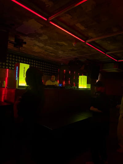 The image depicts the interior of Bar Americas in Guadalajara, Mexico, characterized by its vibrant atmosphere and contemporary design. The scene is illuminated with neon red lights that accentuate the raw texture of the walls. The focus is on the bar area, where patrons are gathered. The bar counter has an illuminated display, with the establishment's name, "bar americas," prominently shown in yellow lettering. Several people are standing at the bar. A surveillance camera is visible near the wall. A decorative panel consisting of circular elements is visible near the bottom of the frame.