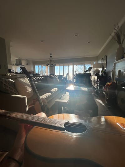 A sunlit living room interior with a large window view. A sofa with patterned cushions and two guitars are in focus. One guitar is held in the foreground, partially obscuring the scene. Sunlight streams through the window, creating strong highlights and shadows. A chandelier hangs in the center of the room. A wheelchair and exercise equipment are visible in the background. The room's neutral palette is accentuated by the warm light.