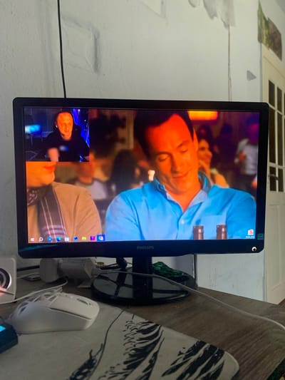 The image shows a computer monitor displaying a video call. On the larger part of the screen, a man in a light blue shirt is visible, seemingly in a casual setting with blurred figures in the background. A smaller window in the lower left corner shows another person, who appears to be the caller, also in a dark outfit and with an orange glow on their face. The monitor is mounted vertically on a wall. To the right of the monitor, a wooden surface is visible with a white computer mouse and some cables. The wall behind the monitor appears to be textured and painted white. There is a window with horizontal panes in the upper left corner of the image. The overall scene suggests a domestic setting, likely indoors, with the video call being the primary focus. The timestamp "30.06.2020" is visible on the top right corner of the monitor.