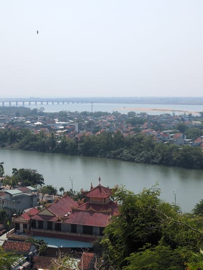 This is an aerial view overlooking the city of Tuy Hòa, Vietnam. The scene contains a river running vertically through the center of the frame. On the river's right bank, a prominent traditional building with a red tiled roof and ornate architecture is visible. The city's skyline with its predominantly red-tiled roofs extends to the left of the river. A long bridge stretches across the horizon. A bird is flying in the pale sky. Trees are visible along the riverbank.