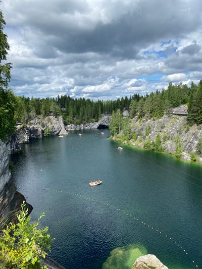 The image depicts the famous Ruskeala Mountain Park in Ruskeala, Russia. The park features a stunning natural landscape of marble canyons filled with clear, emerald-green water. In the foreground, a steep, rocky cliff face covered in lush green vegetation and yellow wildflowers descends into the water. Several small boats, some with people in them, are scattered across the lake. The water is calm, with gentle ripples on the surface. In the background, towering pine trees line the upper edges of the marble cliffs, and a wooden walkway with viewing platforms is visible, allowing visitors to admire the scenery. The sky above is filled with dramatic, gray clouds, interspersed with patches of bright blue sky, suggesting a dynamic weather system. The overall impression is one of natural beauty and recreational activity within a unique geological setting.