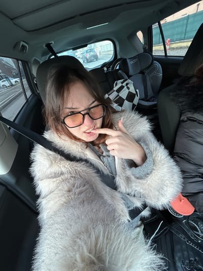 The interior of a vehicle, consistent with a modern SUV or hatchback, serves as the setting for this image, located within Souffelweyersheim, France. A young female is seated in the rear passenger seat on the driver's side, secured by a diagonal seatbelt. She has brown hair and wears large, dark-framed glasses with orange-tinted lenses. Her attire includes a light-colored, possibly faux fur, coat over a grey garment. A visible lesion on her lower lip exhibits redness and dried blood or a scab. Her right index finger is positioned near this lesion, and her mouth is slightly open, revealing upper teeth.
In the rear passenger seat behind the front passenger, a grey child safety seat, partially labeled "bebc...r," is installed. A black and white checkered bag rests on this seat. A second passenger, partially visible on the far right, wears a dark-colored, possibly fur-trimmed, jacket. An orange, possibly silicone, object rests on the floorboard near the second passenger.
Through the vehicle's windows, an urban street scene is visible under daylight conditions. The rear window shows a multi-lane paved road with multiple vehicles in traffic, including a dark-colored SUV directly behind. Buildings line the street, consistent with European architecture. The side window on the driver's side shows a parked white vehicle and additional buildings.