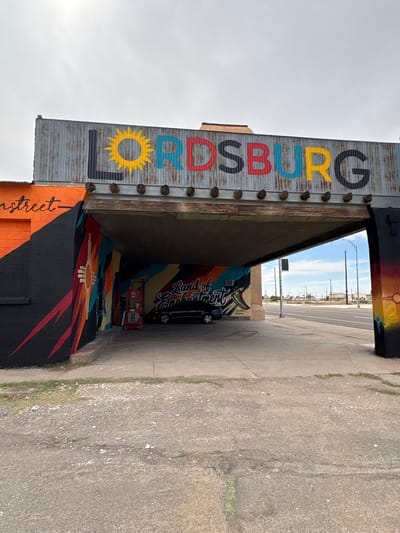 The image depicts a street-level view of an archway entrance in Lordsburg, New Mexico, United States of America. The archway is adorned with large, colorful lettering that spells out "LORDSBURG." The letters are painted in various colors, including black, yellow, blue, and red, against a corrugated metal background. The archway itself is constructed with wood and metal supports.

On the left side of the archway, a building wall is painted with vibrant murals. The mural features a black background with abstract, flame-like shapes in red, orange, and yellow, along with a sun symbol and a stylized representation of the Zia sun symbol. The words "n street" are visible on the orange section of this wall.

The interior of the archway reveals a dimly lit space with a continuation of colorful mural art on the walls. A black SUV is parked inside the archway, and a red, multi-level lift is positioned behind it. The words "Land of Enchantment" are painted in cursive script on the mural inside the archway, accompanied by a drawing of a chili pepper.

To the right of the archway, the mural continues with a sunset motif in yellow, orange, and red, also featuring a sun symbol. Beyond the archway, a two-lane asphalt road is visible, with utility poles and a few distant buildings under a bright, overcast sky. The ground in the foreground is paved with asphalt, showing some cracks and gravel. The overall scene suggests a public art installation or a decorated gateway in the town of Lordsburg.