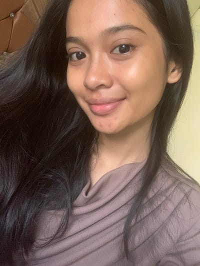 A close-up, selfie-style photograph shows a young woman with long, dark hair. She is looking directly at the camera with a gentle smile. Her skin appears clear and has a slight sheen. She is wearing a muted, brownish-purple shirt with a cowl neck. The background is out of focus, with a textured, light brown surface on the left and a pale yellow surface on the right, possibly indicating an indoor setting. There is no visible indication of the city of Sungai Meriam, Indonesia.