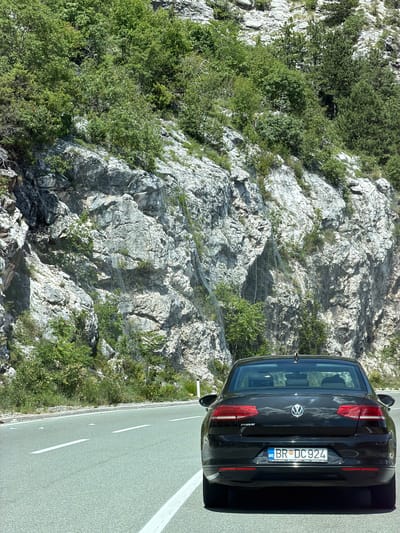 A black Volkswagen Passat is parked on the right side of a two-lane asphalt road, facing away from the camera. The car's license plate reads "BR DC 924." To the left of the car, the road curves slightly, with white lane markings. The background is dominated by a steep, rocky mountainside covered in lush green trees and vegetation. Protective netting is visible on the mountainside, suggesting a potential rockfall hazard. The sky is bright and clear, indicating a sunny day. The car is the primary subject, positioned centrally in the lower portion of the frame. The scene appears to be a scenic mountain road, likely in or near Dobrsko selo, Montenegro, given the mention of the location.