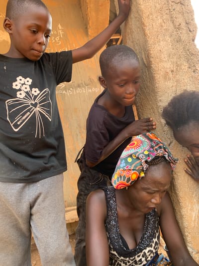 The image depicts three individuals, possibly in Kukum, Nigeria, given the prompt's instruction. One is a woman with dark skin, wearing a leopard-print top with sequined detailing and a colorful headscarf. She is positioned near the top of the frame, lying down. A young boy with dark skin, wearing a black t-shirt, is next to her. Another boy, also with dark skin, wearing a black t-shirt with a white floral design, and grey pants, is lying down near the bottom of the frame. The backdrop appears to be a wall or surface with some writing on it. The scene appears informal and relaxed.