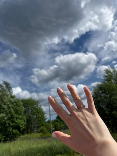 A close-up, slightly angled shot shows a person's left hand, palm facing forward and fingers splayed upwards, in the foreground. The hand has light skin and neatly manicured fingernails painted a pale pink. A thin ring with a striped pattern in gold and possibly another metal is worn on the ring finger. The background is out of focus, featuring a sky with dramatic, dark grey storm clouds interspersed with patches of blue sky and fluffy white clouds. Below the sky, a line of lush green trees and vegetation is visible, with hints of purple flowers in the lower portion of the frame. The overall impression is of a hand reaching towards a dynamic sky, set against a natural, outdoor environment. The image does not provide any specific clues to its location in the city of Chelyabinsk, Russia.