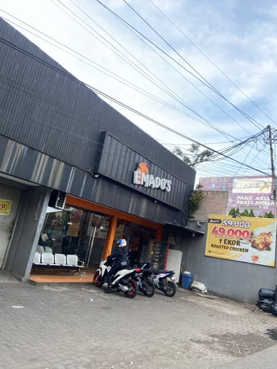 The image shows a storefront of a roasted chicken restaurant called "Emado's" in Kecamatan Margahayu, Indonesia. The storefront is located on a street with a brick walkway, with a large, orange and black sign with the restaurant's name above the entrance. The restaurant is marked with a large, yellow sign outside advertising a roasted chicken for 49,000 rupiah. There is a single customer wearing a blue and white helmet standing on a white motorcycle parked in front of the restaurant. There are several black metal chairs and a small bench in front of the entrance. The image is captured from the perspective of someone standing across the street from the restaurant.