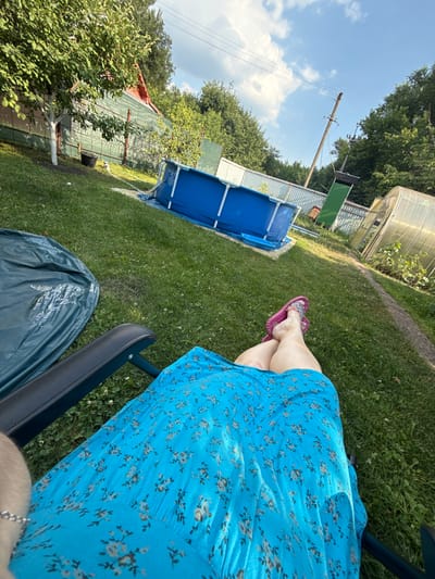 A person, partially visible from the waist down, is sitting in a dark chair in a backyard. They are wearing a blue floral dress and pink flip-flops. The person's legs are crossed, with their feet resting on the grass.
The scene is a sun-drenched backyard with lush green grass. To the left of the person, a blue inflatable swimming pool is set up. In the background, there are trees, a greenhouse, and what appears to be a fence or a building with a corrugated metal roof. A utility pole with wires is visible against a clear blue sky with scattered white clouds.
The time of day appears to be daytime, likely afternoon, given the bright sunlight casting shadows. The weather seems warm and pleasant. The location is suggested to be Nizhne-Chufichevo, Russia, indicated by the presence of a greenhouse and typical backyard elements. The overall mood seems relaxed and peaceful, suggesting a leisurely day spent outdoors. No visible text is present in the image.