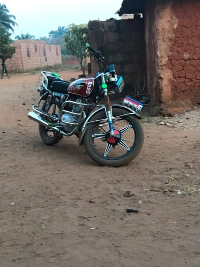 A motorcycle with a distinctive red and black paint scheme, prominently displaying the number "2025," stands parked on a dirt surface. The motorcycle features chrome accents, including a protective cage around the engine and front and rear fenders. A rustic, earthen building stands adjacent to the motorcycle. In the background, partially constructed brick structures are visible amidst scattered trees. The setting suggests a rural or developing area.